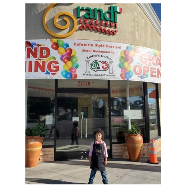 A child in an apron stands outside Grandi Italiani, a restaurant with a "Grand Opening" banner.