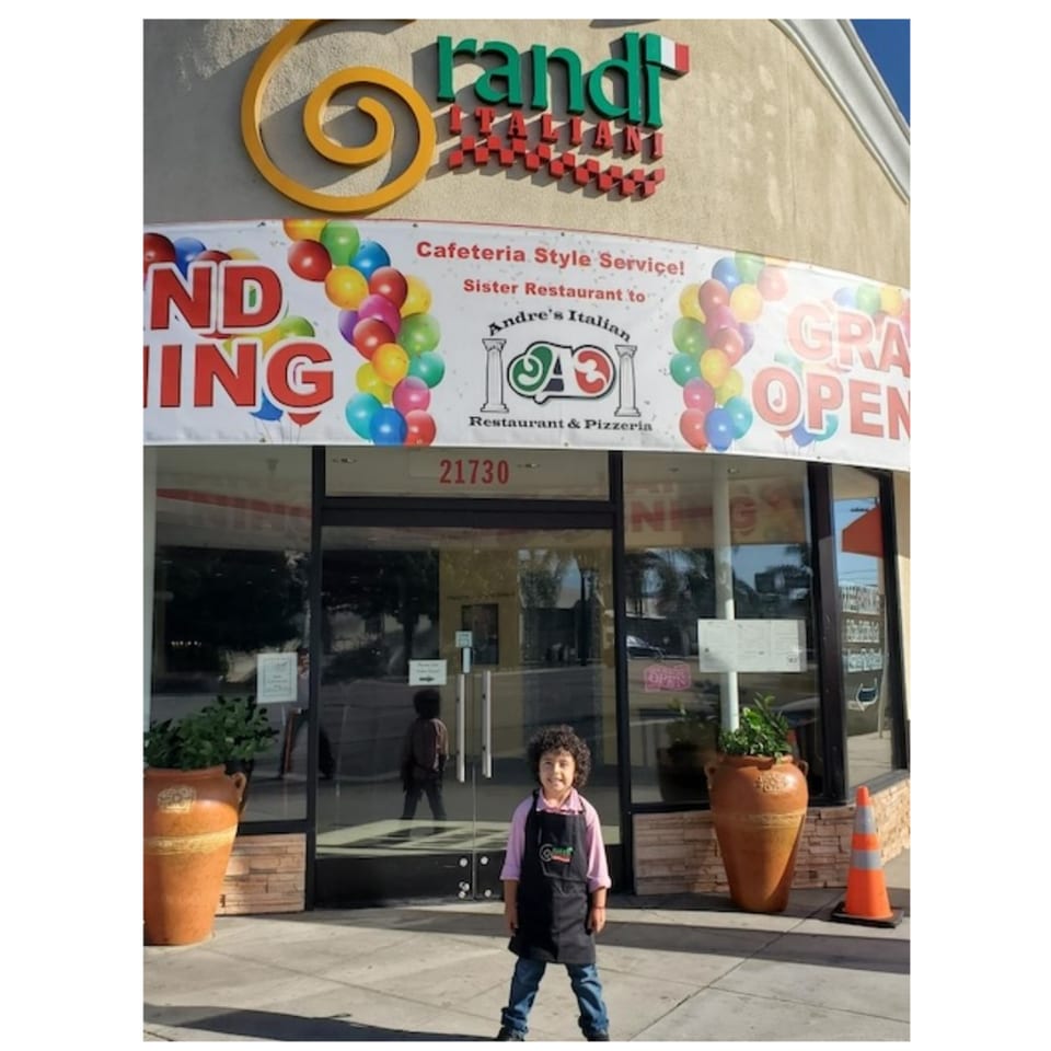A child in an apron stands outside Grandi Italiani, a restaurant with a "Grand Opening" banner.
