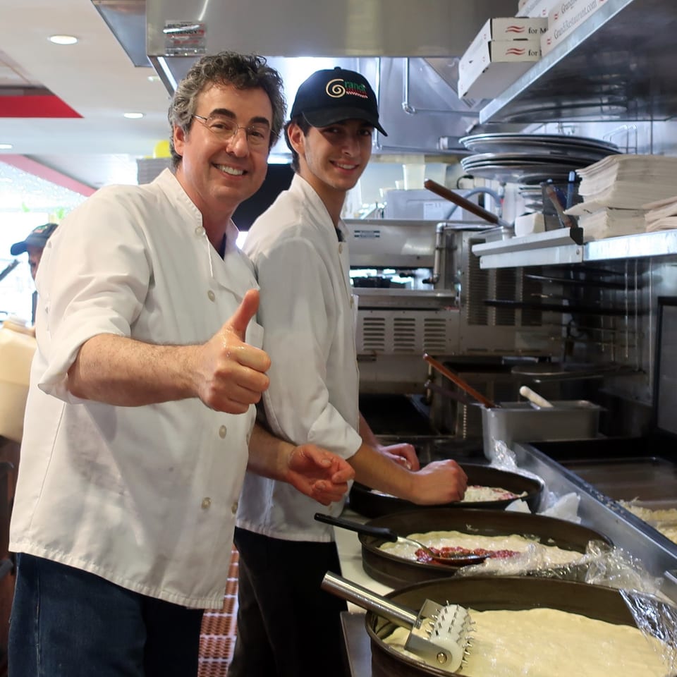 Two Grandi Italiani chefs, one giving a thumbs up, preparing pizzas in a busy kitchen.
